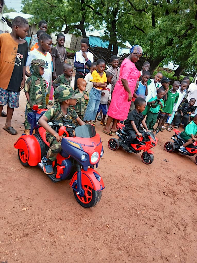 Young learners in camouflage riding toy motorbikes past a crowd